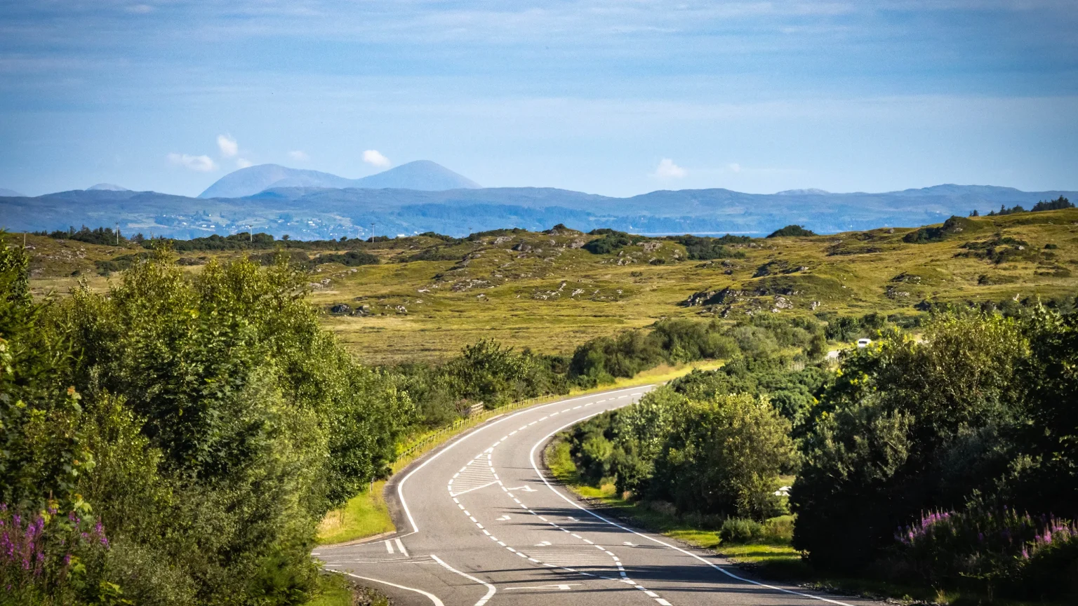 A sunlit, winding road through green hills on the Road to Mallaig
