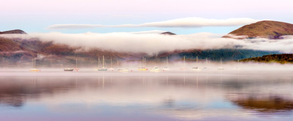 Moored sailing boats on Loch Lomond beneath bands of mist and layered hills.