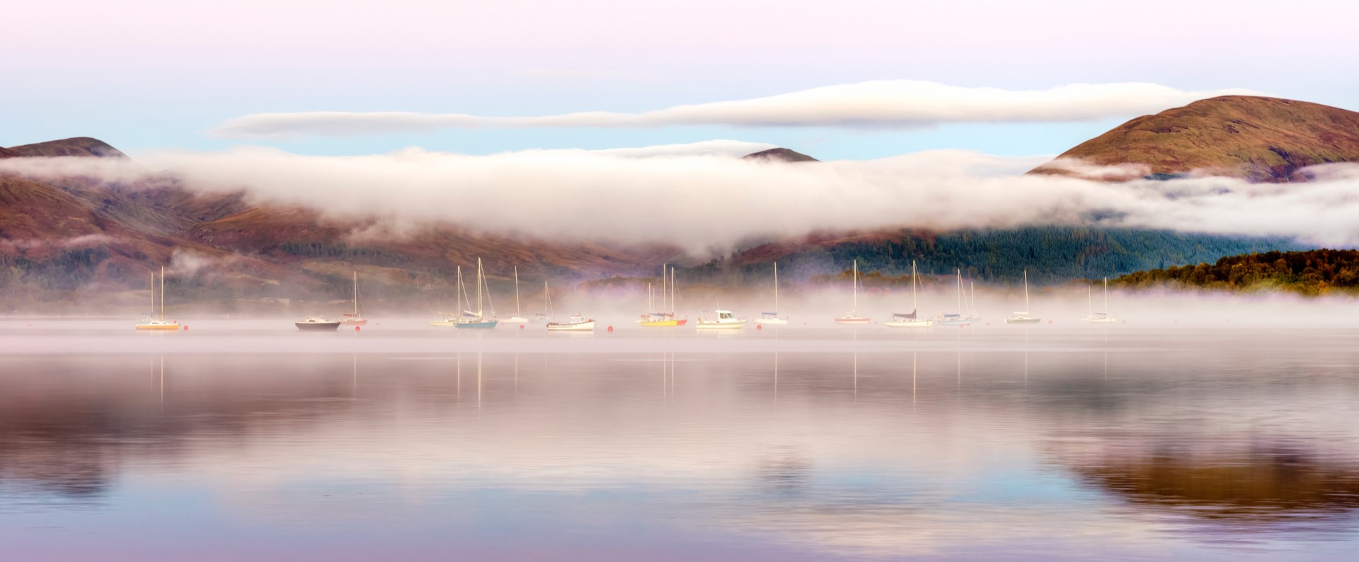 Moored sailing boats on Loch Lomond beneath bands of mist and layered hills.