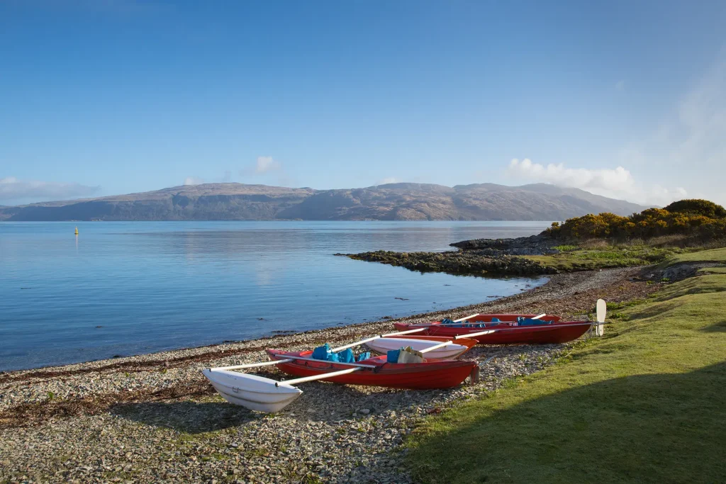 Red and white boats on the pebble shore at Craignure, with calm blue water and hills beyond.