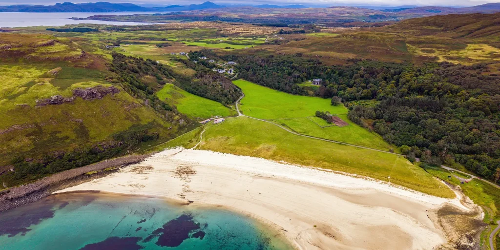 Aerial view of the pale sand and turquoise water of Calgary Bay, backed by green fields and wooded hills.